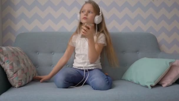 Little girl in headphones is listening to music from smatphone while sitting on sofa at home - Video