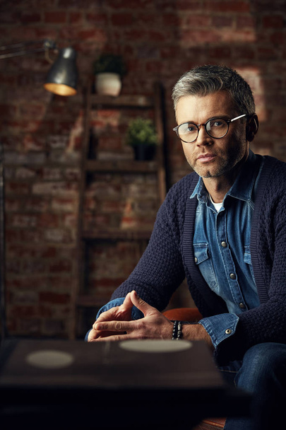 Modern man with gray hair and glasses sitting in studio loft on the sofa and looking at camera - Фото, изображение