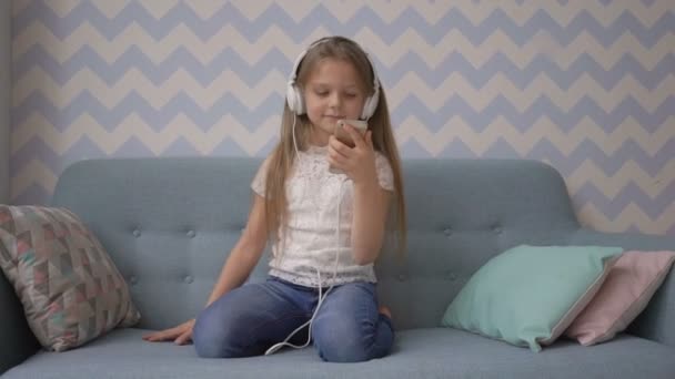 Little girl in headphones is listening to music from smatphone while sitting on sofa at home - Felvétel, videó