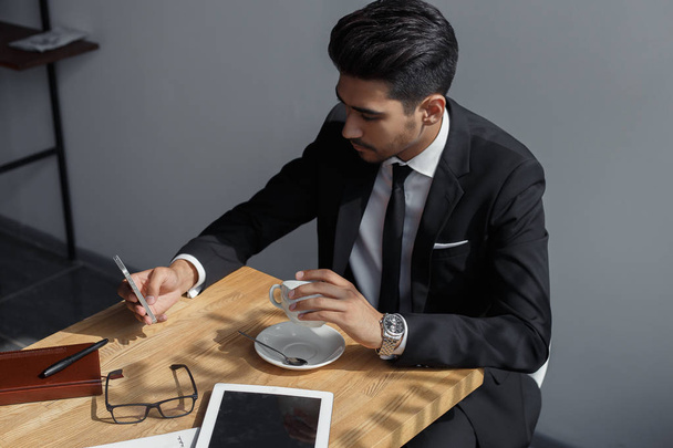 Young businessman holding mobilephone in hand, writing text message, sitting at desk. - Photo, Image