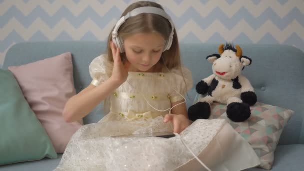 Pretty little girl in headphones is listening to music using a tablet, while sitting on sofa at home - Séquence, vidéo