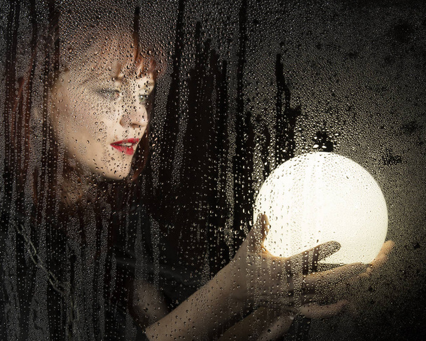 Гладкий портрет женщины-модели, позирующей за прозрачным стеклом, покрытым каплями воды. женщина держит большой светящийся шар
 - Фото, изображение
