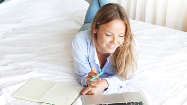 Beautiful caucasian woman working on laptop in the bedroom. Young cute girl talking by the phone and smiling. Nice happy teen lies on the bed and preparing to exams. Sunny morning. - Filmagem, Vídeo