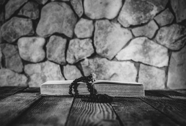 prayer beads with a cross on an open old book on old wooden table on a background of stone walls. selective focus, black & white photo. with space for your text - Foto, Bild