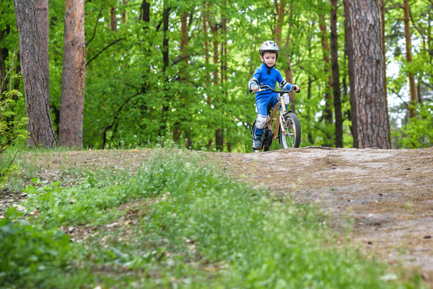 Menino pequeno engraçado feliz na capa de chuva colorida que monta sua primeira bicicleta no dia frio na floresta. Lazer ativo para crianças ao ar livre. conceito de infância despreocupada
 - Foto, Imagem