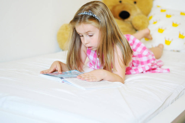 Suloinen pieni tyttö makaa sängyllä ja lukee kirjaa
 - Valokuva, kuva