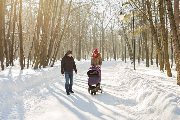 glückliche junge Familie, die im Winter im Park spazieren geht. die Eltern tragen das Baby im Kinderwagen durch den Schnee. - Foto, Bild