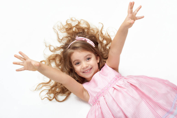 girl child with long hair lie on white and open arms, dressed in pink - Fotografie, Obrázek