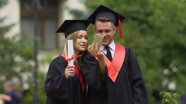 Estudiantes graduados en vestidos académicos tomando selfies después de la ceremonia de graduación
 - Metraje, vídeo