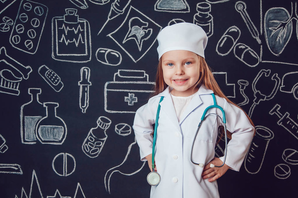 Little girl in doctor costume holding sthetoscope. Dark background with pattern. Her hands on the belt - Фото, зображення