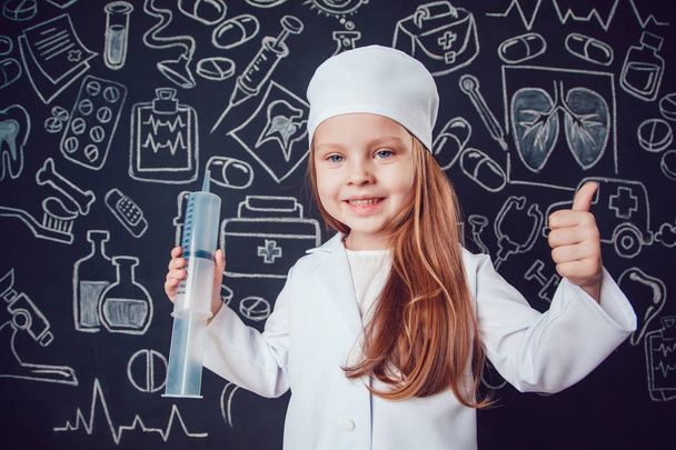 Little girl in doctor costume holding syringe and shows class on dark background with pattern - Foto, afbeelding