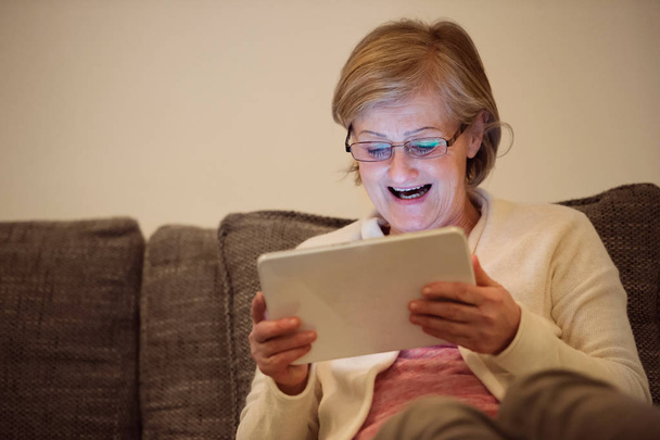Starší žena s tabletem, sedí na gauči v obývacím pokoji - Fotografie, Obrázek