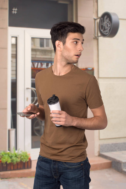 ο νεαρός στέκεται με ένα φλιτζάνι Θέρμο με καφέ και τηλέφωνο - Φωτογραφία, εικόνα