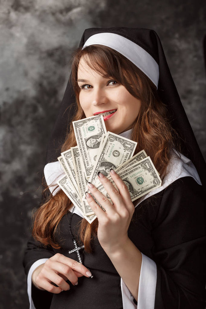 close-up of attractive sexy nun with dark hair holding a dollar bill and a bill lacks teeth on a dark background - Foto, Imagen