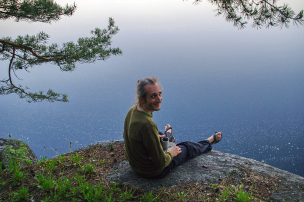 onnellinen nuori mies istuu kallioisella rinteellä järven yläpuolella
 - Valokuva, kuva