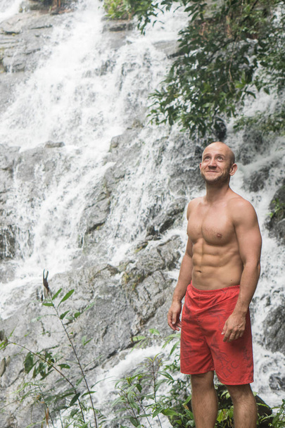 Feliz hombre guapo de pie y mirando hacia arriba cerca de la cascada
 - Foto, imagen