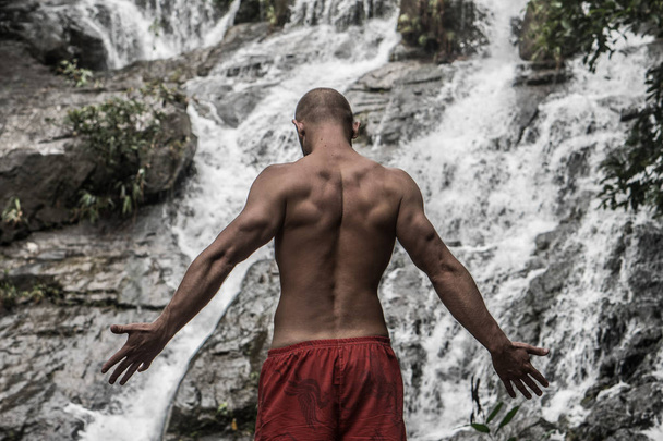 Back view of muscular man standing with raised arms near waterfall - Foto, imagen