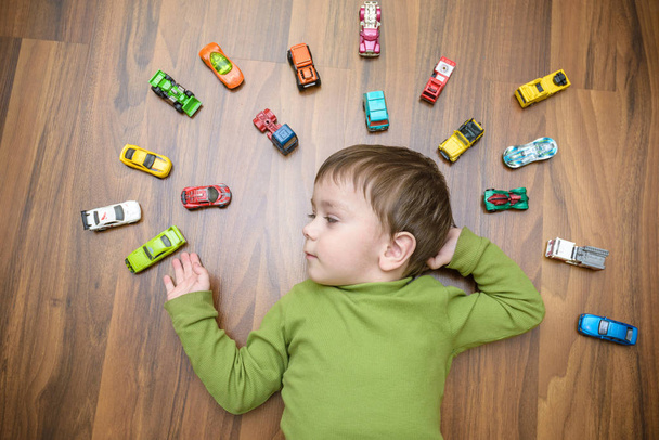 kleines kaukasisches Kind, das drinnen mit vielen Spielzeugautos spielt. Junge im grünen Hemd. fröhliche Vorschulkinder, die Spaß zu Hause oder im Kindergarten haben - Foto, Bild