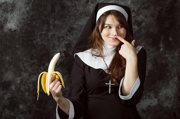 close-up de uma freira sexy segurando uma banana e cartuchos polegar seu nariz em um fundo escuro. Mulher come banana
 - Foto, Imagem
