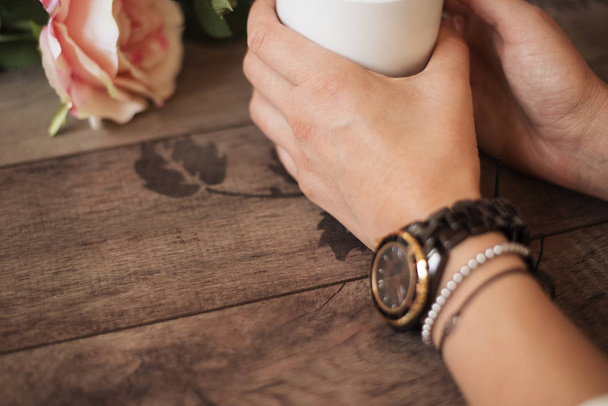 Girl is holding white cup in hands. White mug for woman, gift. Female hands with watch and bracelets holding hot cup of coffee or tea in morning. Selective focus - Photo, Image