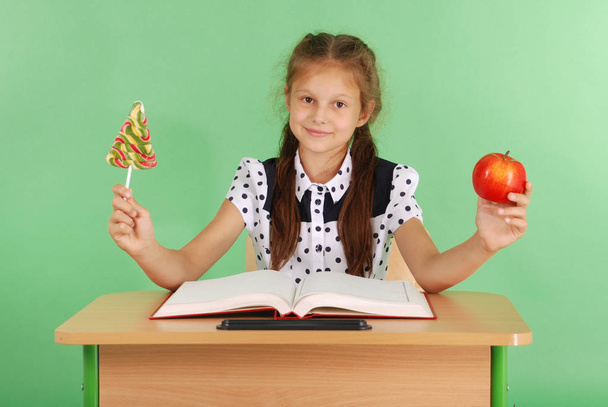 Chica en un uniforme escolar sentado en el escritorio y elegir dulces o una manzana
 - Foto, imagen