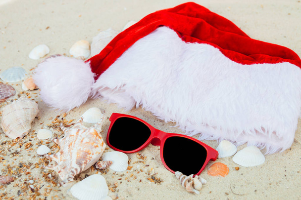 Christmas hat and red sunglasses on the beach. Santa   eyeglasses  the sand near shells. Holiday. New year vacation. Copy space. Frame. Top view. - Photo, Image
