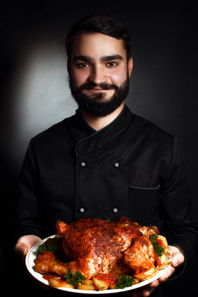 Επαγγελματίας σεφ με μια γενειάδα με μαύρες στολές με μαγειρεμένο κοτόπουλο στα χέρια του - Φωτογραφία, εικόνα