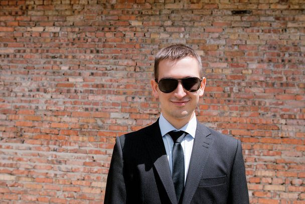 jovem em terno preto, gravata e óculos de sol pretos no fundo da parede de tijolo
  - Foto, Imagem