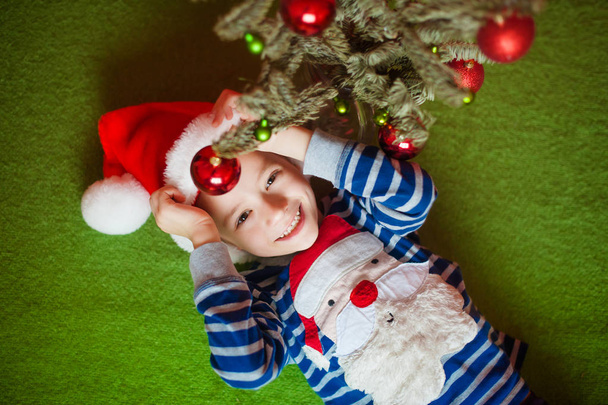 Happy little boy is lies near fir-tree. New Years holidays.  in a striped T-shirt with Santa Claus - 写真・画像