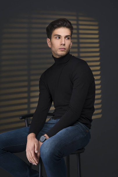 stylischer junger Mann im schwarzen Pullover sitzt neben Jalousien und Denkern - Foto, Bild
