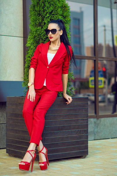 Retrato de una joven hermosa mujer de negocios de cabello oscuro con gafas de sol y traje rojo brillante sentada en el edificio de oficinas con ventanas espejadas
 - Foto, Imagen