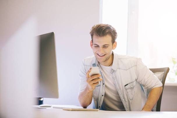 Smiling man talking on mobile phone while using laptop computer at desk in study. - 写真・画像