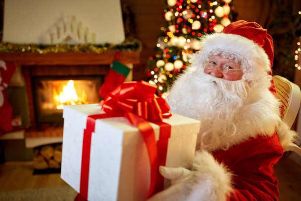 Santa Claus sitting by the fireplace with big present - Foto, afbeelding