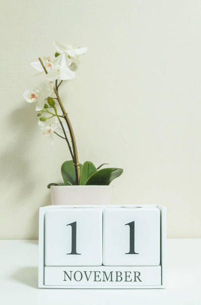 Closeup witte houten kalender met zwarte 11 november word met witte orchidee bloem op witte houten bureau en crème kleur behang in de kamer getextureerde achtergrond, selectieve aandacht op de kalender - Foto, afbeelding