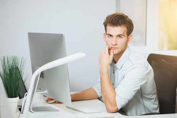 A young programmer is looking at the camera. The graphic designer is thinking about the future projects. The young guy is sitting at the computer in the office with the windows behind his back - Φωτογραφία, εικόνα