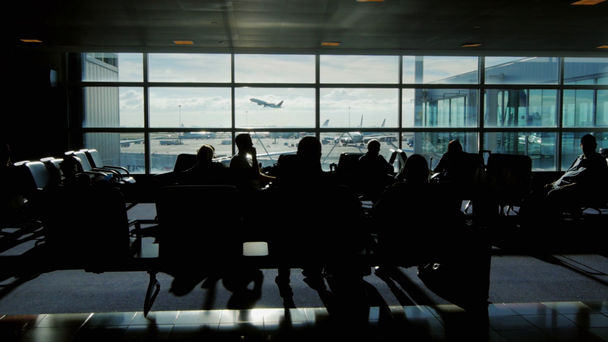 Ludzie czekają na lot w terminalu lotniska w słoneczny dzień. Za oknem samolot startuje. Można zobaczyć sylwetki ludzi, nie rozpoznawalne twarze - Materiał filmowy, wideo