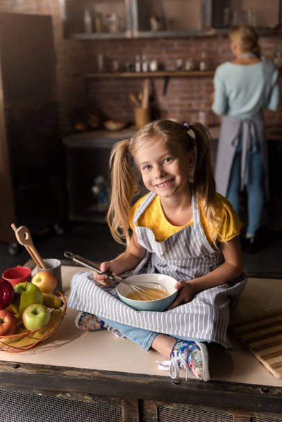 Cheerful smiling girl mixing eggs while sitting on the table - Valokuva, kuva
