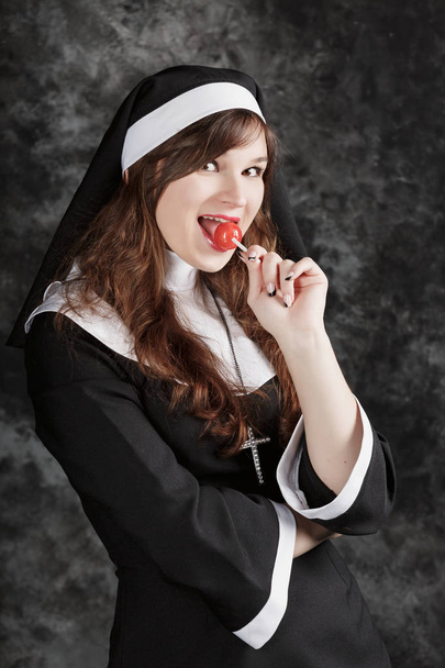 karanlık bir arka plan üzerinde kırmızı bir şeker yalama çorap genç seksi rahibe. Bonbon yemek çekici kadın sahiptir - Fotoğraf, Görsel
