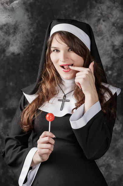 young sexy stockings nun holding a red bonbon on a dark background.attractive brunette girl with candy licking finger. - Photo, Image
