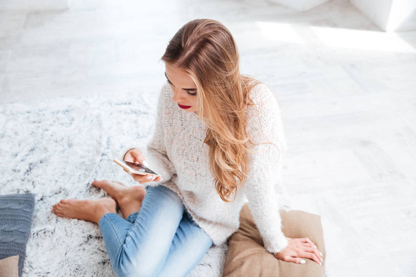 Young woman using mobile phone while sitting on the carpet - Photo, Image