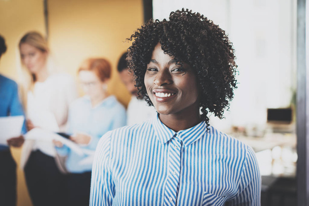 Portrait of pretty african american business woman with afro smiling at the camera.Coworking team on background in modern office. Horizontal,blurred. - Zdjęcie, obraz