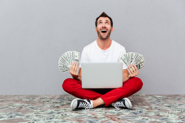 Man holding banknotes and sitting on the floor with laptop - Photo, Image