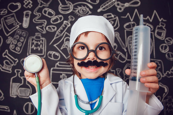 Happy little boy in doctor costum holding syringe and sthetoscope on dark background with pattern. The child has mustache, glasses - Фото, изображение