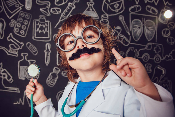 Happy little boy in doctor costume holding sthetoscope on dark background with pattern. Lifts thumbs up. The child has mustache, glasses - Fotoğraf, Görsel
