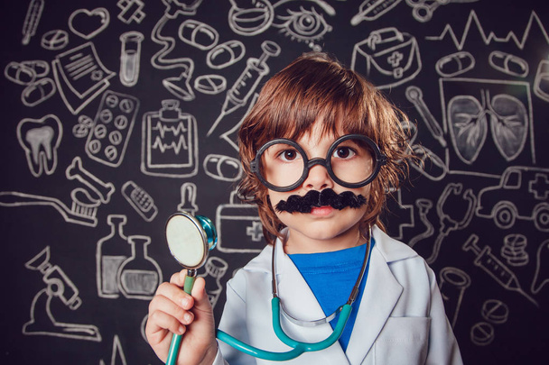 Счастливый маленький мальчик в костюме доктора держит стетоскоп на темном фоне с рисунком. У ребенка есть усы, очки
 - Фото, изображение