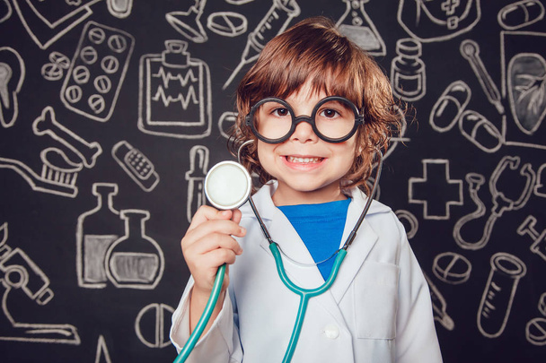 Happy little boy in doctor costume holding sthetoscope on dark background with pattern. The child has glasses - Foto, afbeelding