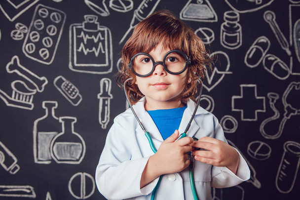 Happy little boy in doctor costume holding sthetoscope on dark background with pattern. The child has glasses - Φωτογραφία, εικόνα