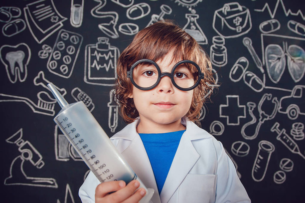 Happy little boy in doctor costume holding syringe on dark background with pattern. The child has glasses - Foto, Imagem
