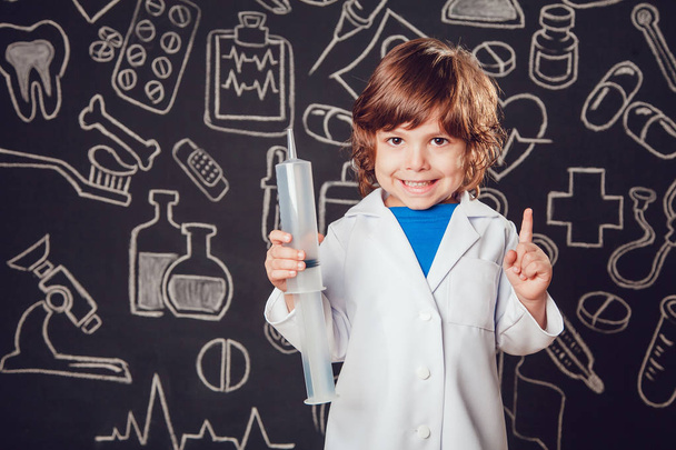 Happy little boy in doctor costume holding syringe on dark background with pattern. Lifts thumbs up. - Photo, Image