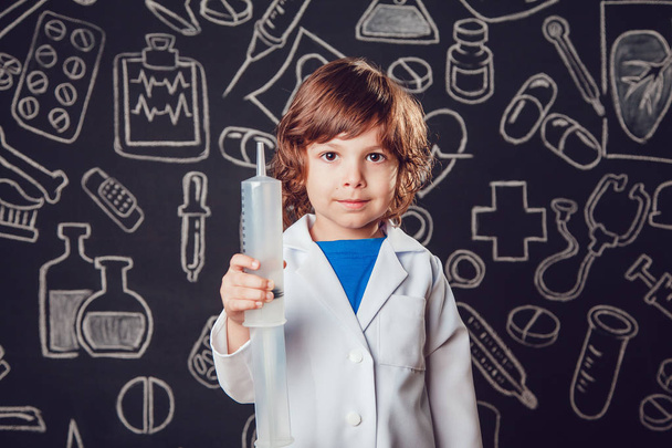 Happy little boy in doctor costume holding syringe on dark background with pattern. - Foto, Bild